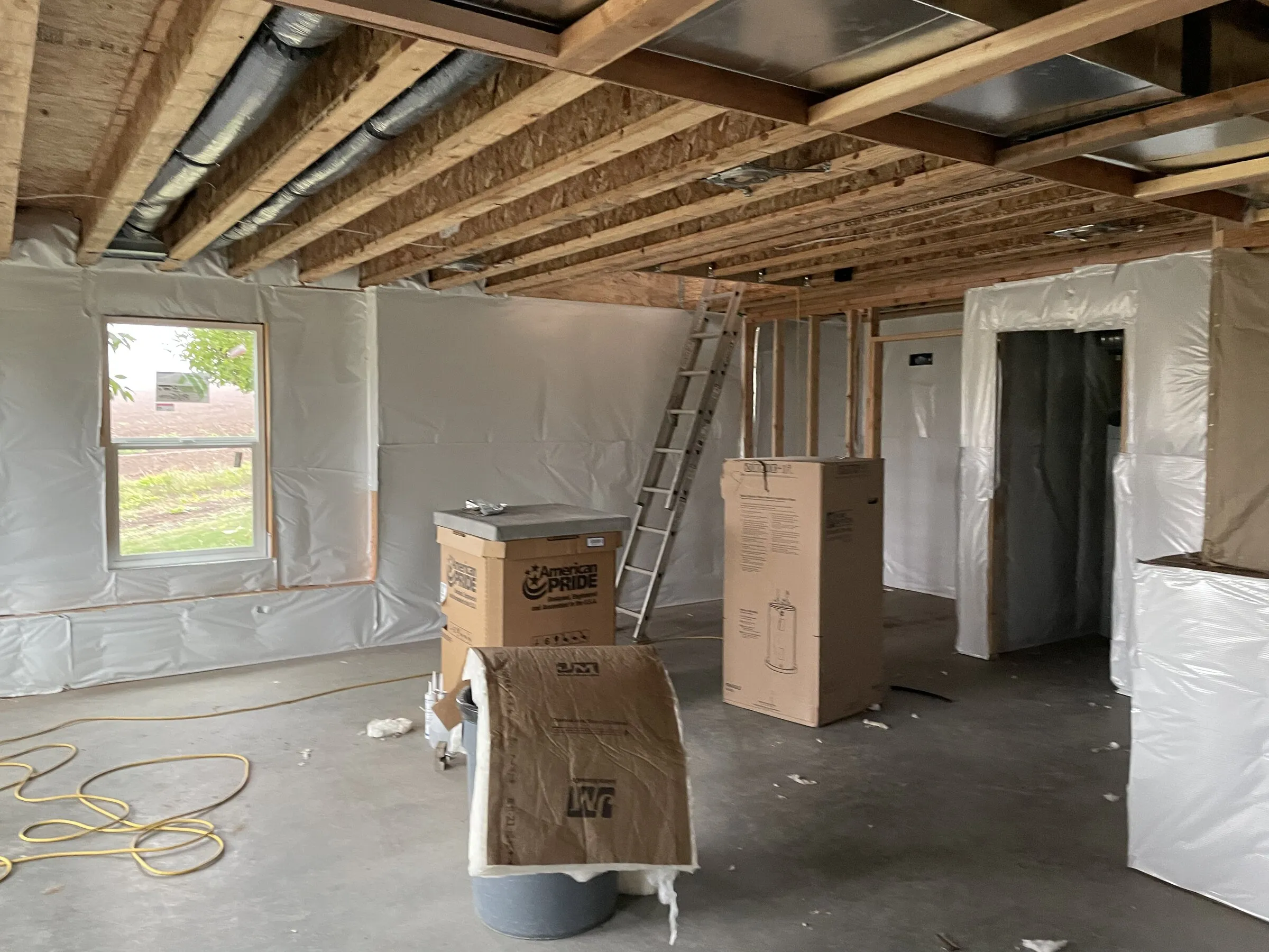 Lower-floor room with the ceiling joists and ductwork exposed overhead and plastic vapor barrier on the walls before drywall